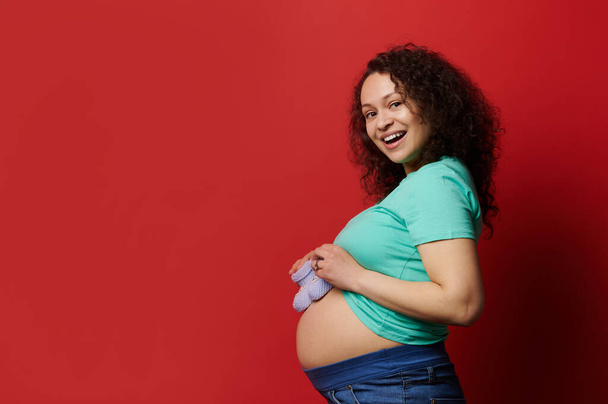 Aantrekkelijk mooie krullende etnische zwangere vrouw met blote buik, poseren met schattige blauwe gebreide baby laarsjes, glimlachen met vrolijke tanden glimlach, geïsoleerd op rode achtergrond. Advertentie kopiëren - Foto, afbeelding