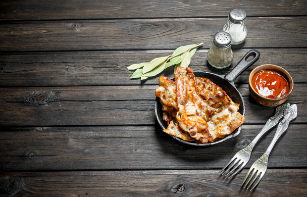 Fried bacon in a pan with tomato sauce and spices. On a wooden background. - Photo, Image