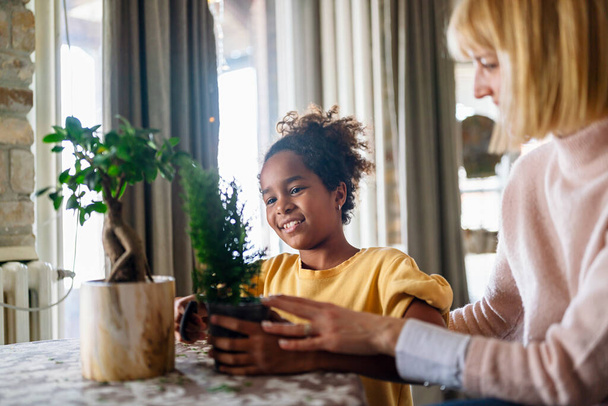 Happy caucasian mother and her adopted african american daughter taking care of plants indoors. Family hobby concept. - Foto, imagen