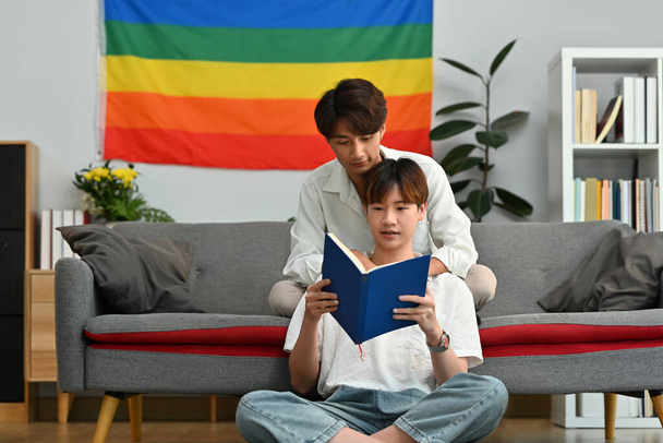 Happy gay couple reading book on sofa at home, spending time together on weekend. LGBTQ people lifestyle and love emotion. - Photo, Image