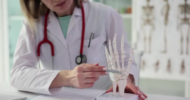 Woman doctor in uniform points pen to skeleton hand sitting at table in clinic office. Professional in field of medicine presents skeleton of arm part slow motion - Footage, Video