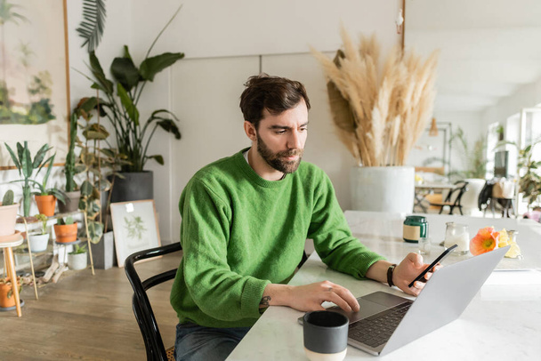 freelancer en puente verde sosteniendo smartphone y usando laptop mientras trabaja cerca de la taza de café - Foto, imagen