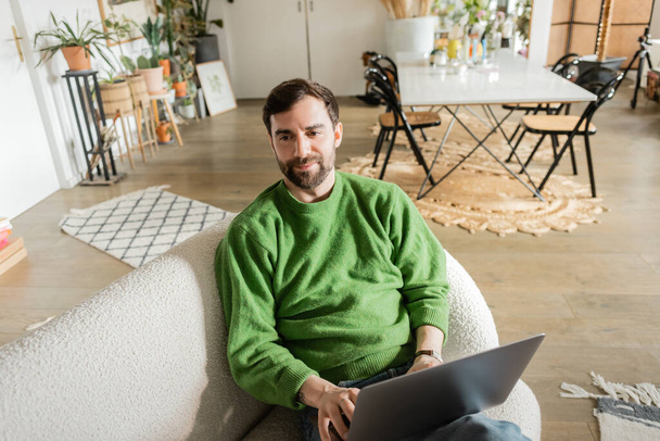 Χαρούμενος και γενειοφόρος ελεύθερος επαγγελματίας σε πράσινο jumper χρησιμοποιώντας φορητό υπολογιστή και κοιτάζοντας μακριά, απομακρυσμένη εργασία - Φωτογραφία, εικόνα