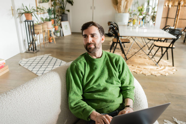 Bearded and brunette freelancer in casual green jumper looking away while using laptop at home  - Photo, Image