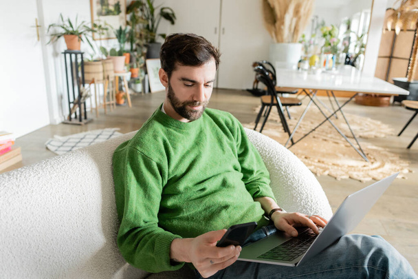 Bearded freelancer in green jumper and jeans using smartphone while working on laptop, remote work - Foto, Imagen