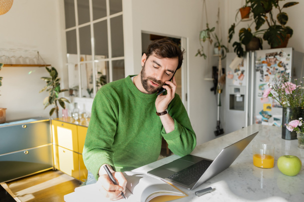 Bearded freelancer in green jumper talking on smartphone and writing on notebook, remote work - Photo, Image
