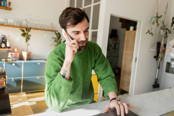 Bärtiger und tätowierter Freiberufler im Pullover spricht auf dem Smartphone, während er zu Hause am Laptop arbeitet  - Foto, Bild