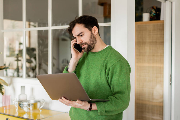 Focused freelancer in green jumper holding laptop and talking on smartphone at home  - Photo, Image