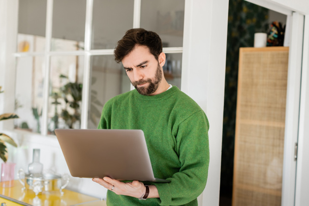 Yeşil kazaklı, sakallı, evde çalışırken laptop kullanan, uzaktan kumandalı bir serbest çalışan. - Fotoğraf, Görsel