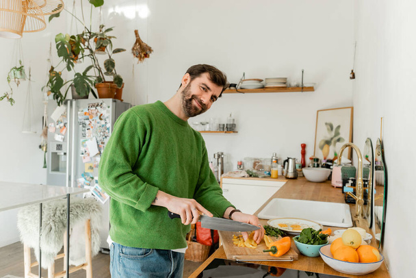 Lächelnder bärtiger Mann in Pullover und Jeans schneidet frischen Pfeffer, während er in der Küche frühstückt  - Foto, Bild