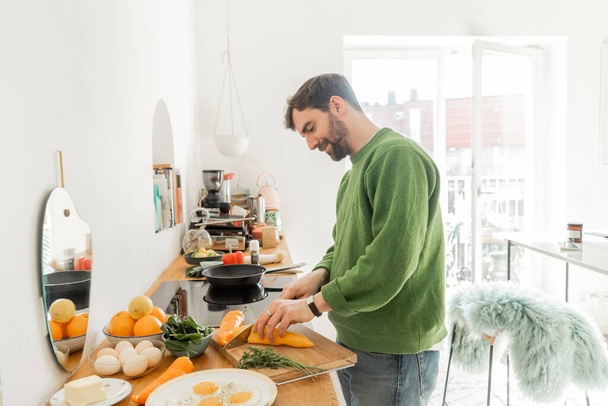 hymyilevä mies vihreässä hyppääjä leikkaamalla tuoretta pippuria, kun ruoanlaitto aamiainen moderni keittiö  - Valokuva, kuva