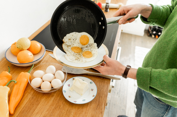 Zugeschnittene Ansicht des Mannes mit Pfanne mit Spiegeleiern in der Nähe des Tellers beim Kochen Frühstück am Morgen  - Foto, Bild