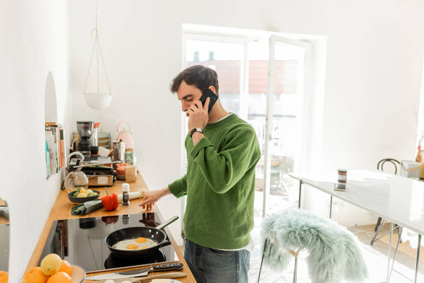 Brunette man in jumper and jeans talking on smartphone while cooking breakfast in kitchen at home - Photo, Image