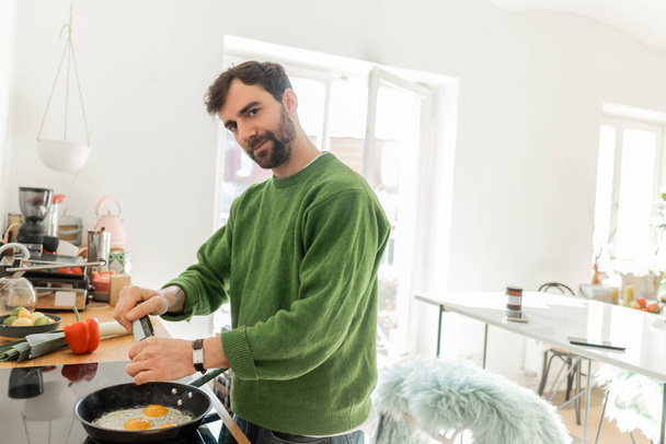 panierter Mann in grünem Pullover blickt in die Kamera, während er Spiegeleier zum Frühstück kocht  - Foto, Bild