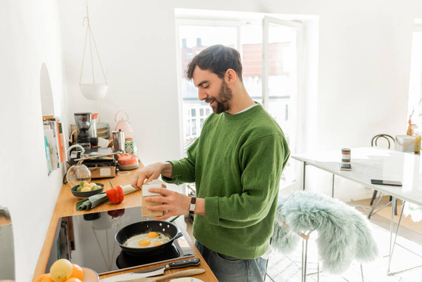 bärtiger Mann im grünen Pullover würzt Spiegeleier beim morgendlichen Frühstück  - Foto, Bild