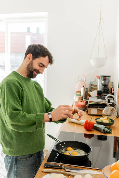 Веселый бородатый мужчина наливает яйцо на сковородку во время приготовления пищи рядом размытые свежие продукты на рабочем столе - Фото, изображение