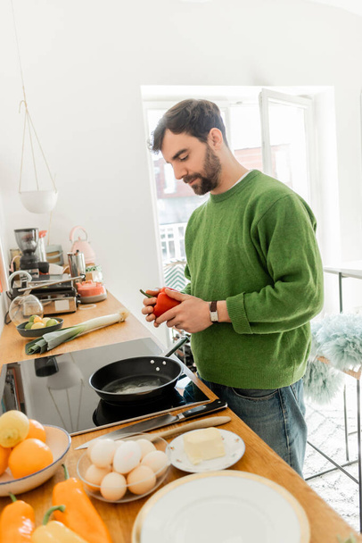 Бородатый мужчина в свитере держит свежий перец возле сковороды, размытые яйца и масло на столешнице - Фото, изображение