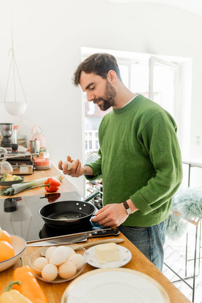 Бородатый мужчина в джемпере держит яйцо возле сковороды, масло и свежие продукты на рабочем столе  - Фото, изображение
