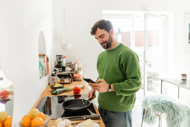 Бородатий чоловік смартфон і тримає сковороду біля яєць, свіжу їжу та масло на робочому столі - Фото, зображення