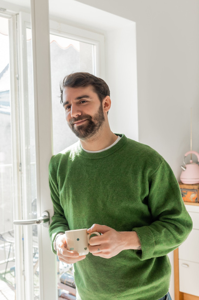 Positive bearded and brunette man in green jumper holding cup of coffee while standing near window - Photo, Image