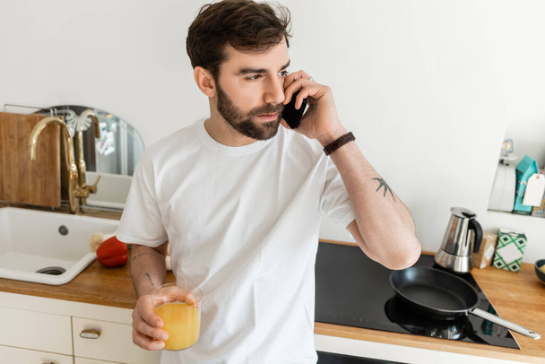 Tetovált szakállas férfi fehér pólóban beszél okostelefonon és kezében üveg friss narancslé  - Fotó, kép
