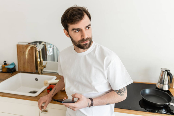 Homme barbu et tatoué en t-shirt blanc regardant loin et tenant smartphone  - Photo, image