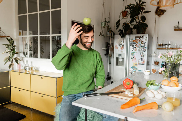 Fröhlicher und bärtiger Mann im grünen Pullover balanciert mit Apfel auf dem Kopf in moderner Küche  - Foto, Bild