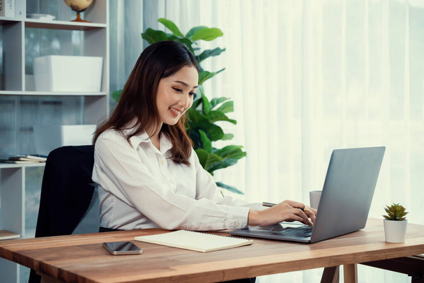 Junge asiatische begeisterte Geschäftsfrau am modernen Schreibtisch mit Laptop, um zu arbeiten und Notizen zu schreiben. Fleißige und attraktive Bürokauffrau arbeitet in ihrem Büro am Computer-Notizbuch. - Foto, Bild