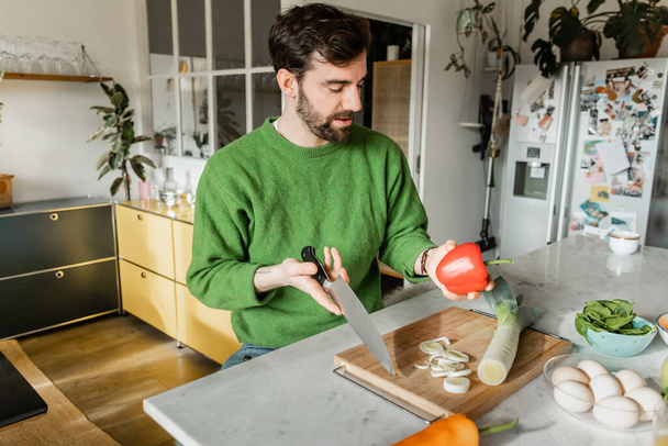 Bearded tattooed man in green jumper holding knife and bell pepper while cooking in modern kitchen - Фото, зображення