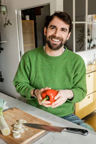 自宅でキッチンで新鮮なベルペッパーを保持する緑のジャンパーの肯定的なひげ付き男  - 写真・画像
