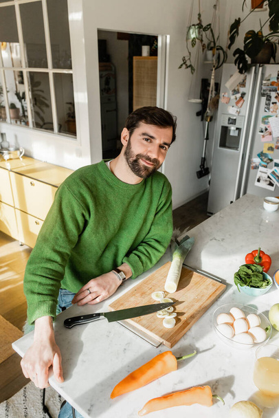 Positiver Mann im Pullover blickt in die Kamera, während er zu Hause in der modernen Küche neben frischen Lebensmitteln sitzt  - Foto, Bild