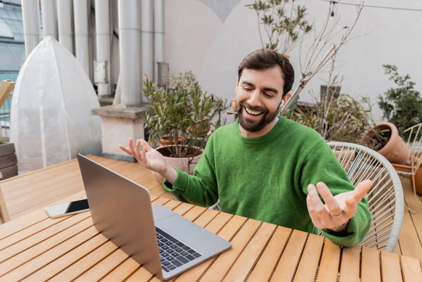 Veselý vousatý muž ve sluchátkách s videohovory na notebooku při práci na střešní terase  - Fotografie, Obrázek