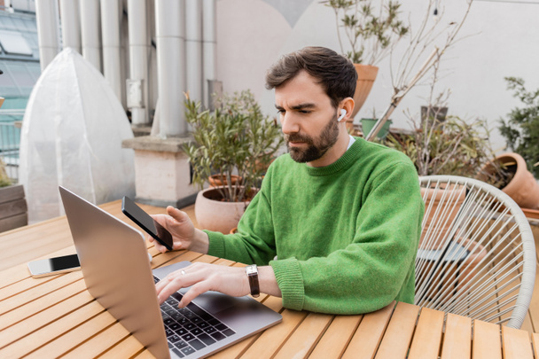Baard ondernemer in oortelefoon en groene jumper holding smartphone en met behulp van laptop in het terras van het huis  - Foto, afbeelding