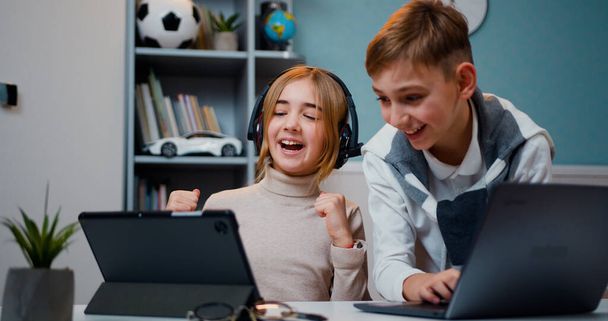 Caucasian boy and girl playing game on laptop and tablet device together with smile face at home in games room. Jojful children sitting the table play online video games and browsing social media in - Fotografie, Obrázek
