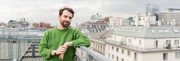 hombre tatuado en jersey verde mirando hacia otro lado mientras está de pie en la terraza de la azotea en Viena, pancarta - Foto, imagen