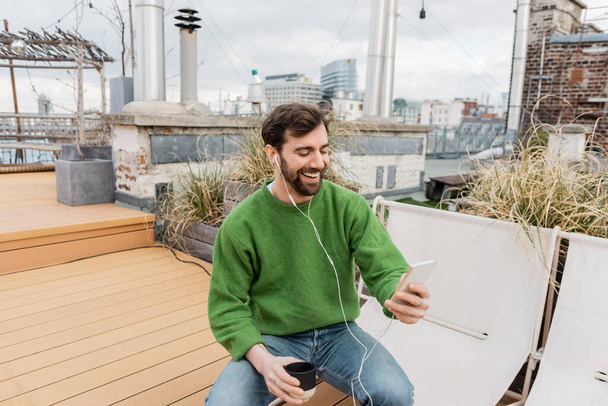 Cheerful man in earphones holding cup of coffee while spending time on rooftop terrace in Vienna - Photo, Image