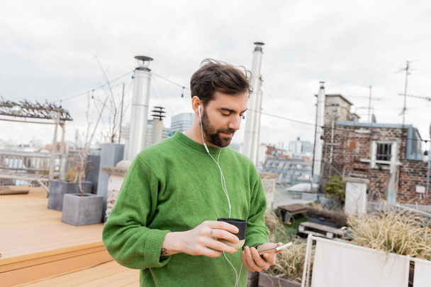 handsome man listening music in earphones and holding cup of coffee on rooftop in Vienna, Austria - Photo, Image