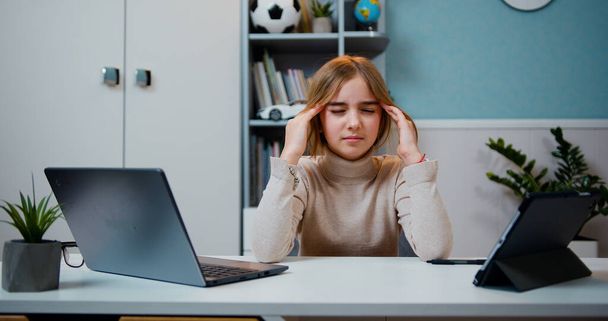 Tired preteen girl sitting by table with headache massaging her temples with circular motion after a hard day on background bookcase at home - Фото, зображення