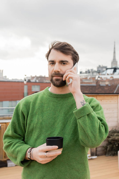 man in jumper talking on smartphone and holding cup of coffee on rooftop in Vienna, Austria - Photo, Image