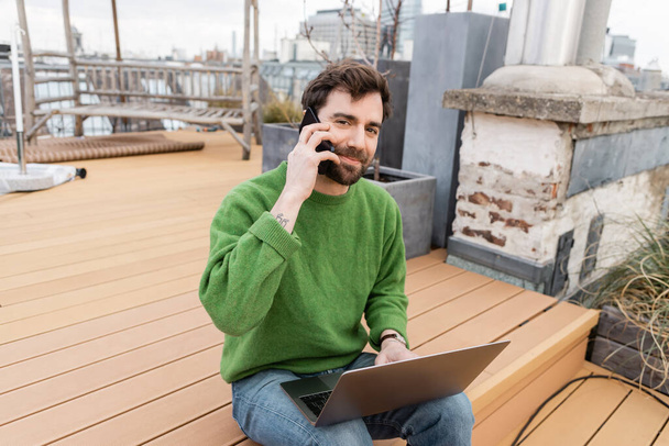 бородатый мужчина разговаривает на смартфоне и работает на ноутбуке на террасе на крыше в Вене, Австрия - Фото, изображение