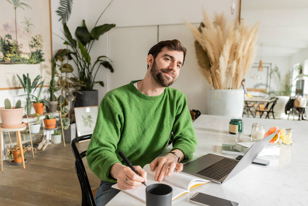 Χαμογελώντας και γενειοφόρος ελεύθερος επαγγελματίας στο πράσινο jumper γράφοντας στο σημειωματάριο κοντά συσκευές στο τραπέζι - Φωτογραφία, εικόνα