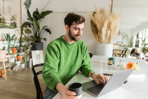 happy freelancer in jumper using laptop and holding cup of coffee while working at home - Photo, Image