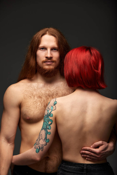Сексуальная полуобнаженная пара влюблена. Привлекательный мужчина с длинными волосами и бородой смотрит в камеру, женщина с рыжими волосами и цветной татуировкой нежно обнимает своего мужчину - Фото, изображение