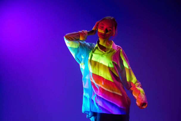 Portrait of young girl with multicolored neon filter reflection on body, posing, dancing against purple background. Concept of art, modern style, cyberpunk, futurism and creativity - Fotografie, Obrázek