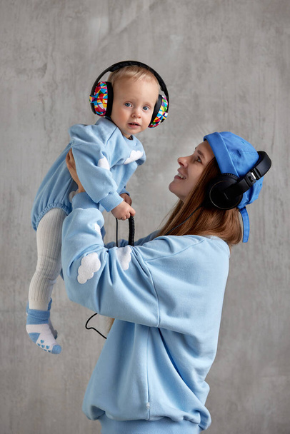 A happy young mother hugs her smiling baby and lifts him up in her arms. Baby and mom in blue clothes and DJ headphones on their heads - Фото, изображение