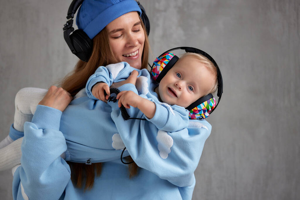 A happy young mother hugs her smiling baby and lifts him up in her arms. Baby and mom in blue clothes and DJ headphones on their heads - Fotoğraf, Görsel