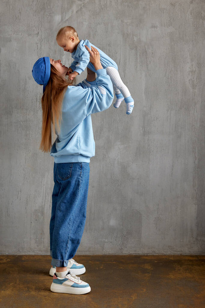 Porträt einer glücklichen jungen Mutter mit Säugling auf dem Arm. Mama hebt ihr süßes Baby auf und betrachtet es von unten nach oben. Baby und Mutter in den gleichen blauen Kleidern. Familienliebe und Harmonie. - Foto, Bild