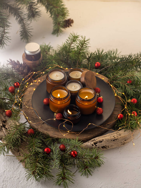 Soy candles burn in glass jars. Comfort at home. Candle in a brown jar. Scent and light. Scented handmade candle. Aroma therapy. Christmas tree and winter mood. Cozy decor. Festive garland decoration - 写真・画像
