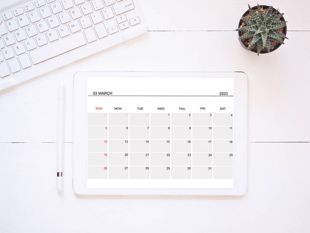 Concepto de negocio y educación. Aplicación de calendario con planificación. Vista superior, Tablet PC que muestra la página del calendario de marzo 2023 y lápiz lápiz óptico, teclado y cactus en el lugar de trabajo blanco - Foto, Imagen
