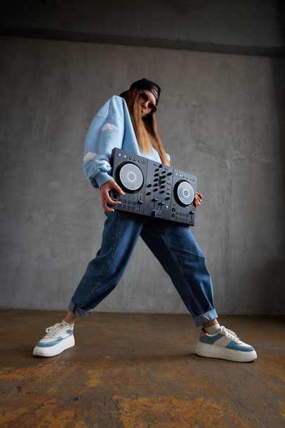 A young pretty long haired hip hop girl in a blue sweater, loose jeans and a black baseball cap holds a dj console in her hands and dances vigorously. Music, fun and creativity - Valokuva, kuva